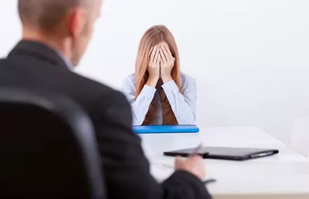Person at a desk with hands covering face, another sitting nearby.