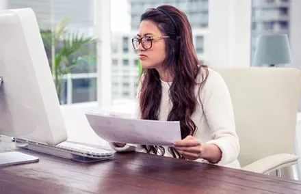 Person wearing glasses, focused on a document while sitting at a desk in front of a computer.