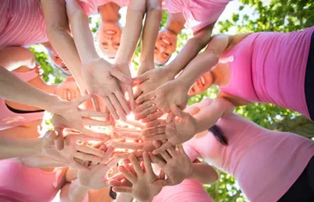 Group of people in pink tops with hands joined in a circle, sunlight shining through.