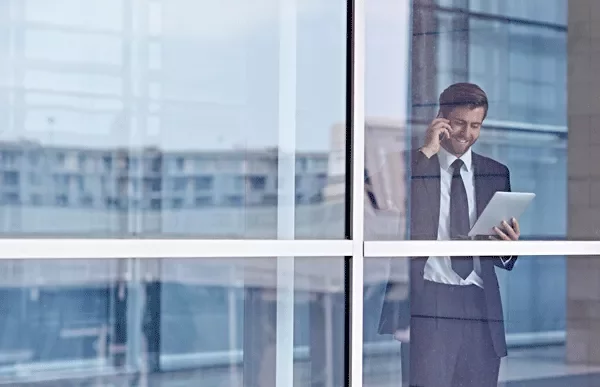 Person in office attire on a phone call while holding a tablet, viewed through a window.