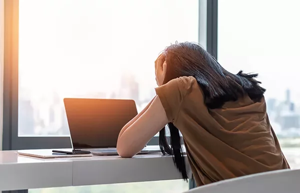 Person resting head on desk near a laptop in a bright room.